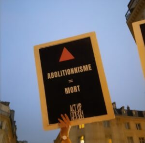 Image d'une pancarte de ACT-UP PARIS en manifestation, sur laquelle est écrite : "ABOLITIONNISME = MORT". Un triangle rose est présent au-dessus du mot "ABOLITIONNISME" et la pancarte a un fond noir, le slogan est en blanc.
