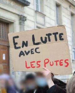 Image d'un.e manifestant.e anonyme brandissant une pancarte qui indique : "EN LUTTE AVEC LES PUTES". La partie "EN LUTTE AVEC" est écrit avec de la peinture noire et "LES PUTES" en rouge. 