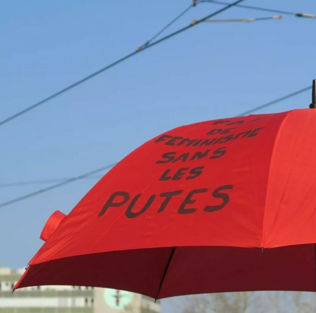 Image d'un parapluie rouge (symbole de la lutte pour les droits des travailleureuses du sexe). Sur ce parapluie rouge est noté : "PAS DE FÉMINISME SANS LES PUTES" à la peinture noire.
